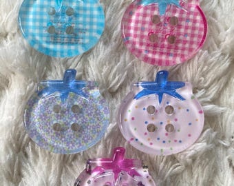May include: Five decorative strawberry-shaped buttons with blue stems. The buttons feature various designs, including gingham, confetti, and floral patterns. The buttons are made of clear plastic.