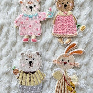 Pode incluir: Quatro patches bordados com personagens de animais. Um veado com chifres rosa e um vestido rosa segura um cupcake. Um urso com um vestido rosa segura uma flor. Um urso com uma camisa listrada amarela segura uma cenoura. Um coelho com um vestido listrado.