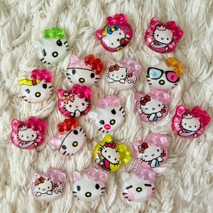May include: A selection of Hello Kitty resin embellishments. The charms depict the character in different poses, with pink bows and accessories. The embellishments are white, pink, yellow, and green, and are scattered on a white, textured surface.