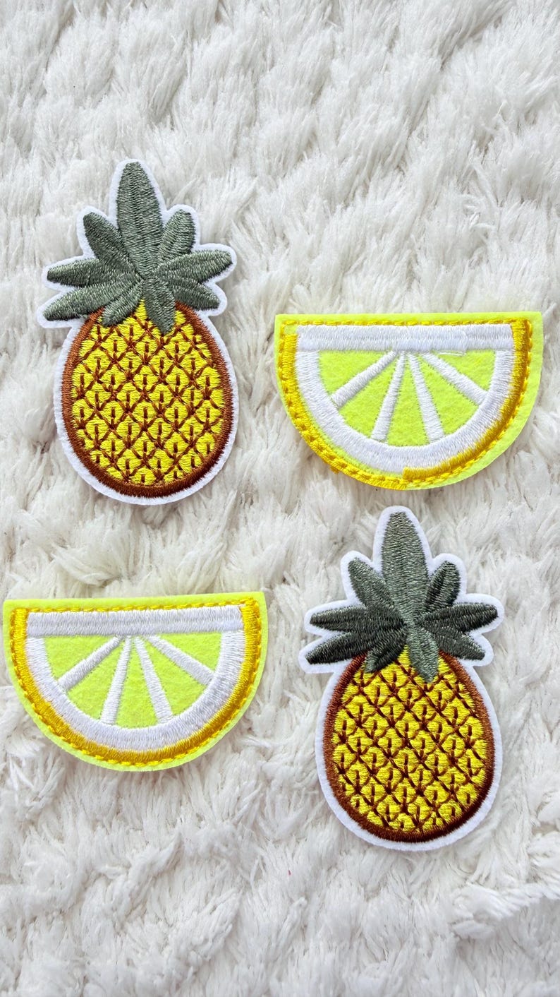 May include: Four embroidered fruit patches. Two pineapple patches with yellow and brown details and green tops. Two lemon slice patches with yellow and white details and yellow borders. All patches are on a white, textured background.