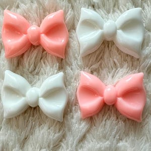 Può includere: Quattro ornamenti lucidi a forma di fiocco, rosa e bianchi. I fiocchi sono su una superficie bianca e soffice. I fiocchi hanno un design arrotondato e tridimensionale, ideali per il fai-da-te o la decorazione.