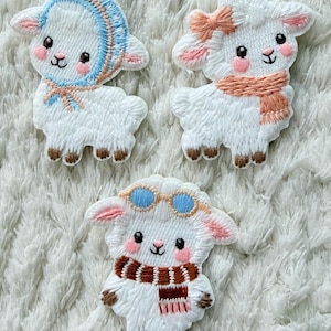 Op de afbeelding: Drie geborduurde schapenpatches. Eén schaap draagt een blauwe muts, een ander een roze strik en sjaal, en de derde een bruine en rode sjaal en een blauwe bril. De schapen zijn wit met roze wangen en bruine hoeven.