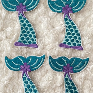 May include: Four teal and purple mermaid tail iron-on patches. Each patch features a teal tail with purple scales and a purple flower.