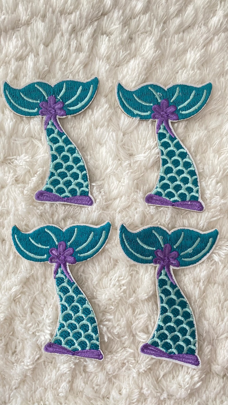 May include: Four teal and purple mermaid tail iron-on patches. Each patch features a teal tail with purple scales and a purple flower.