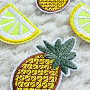 May include: Embroidered patches with pineapple and lemon slice designs. The pineapple patches have a yellow body with brown detailing and green leaves. The lemon slices are yellow with white details. These patches are suitable for adding to clothing or accessories.