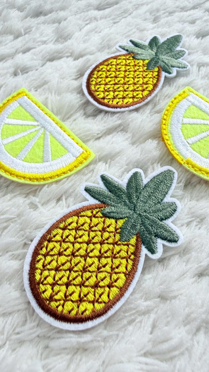 May include: Embroidered patches with pineapple and lemon slice designs. The pineapple patches have a yellow body with brown detailing and green leaves. The lemon slices are yellow with white details. These patches are suitable for adding to clothing or accessories.
