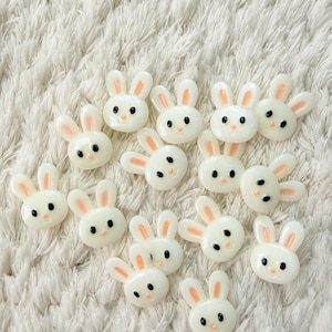 Puede incluir: Una colección de adornos blancos con forma de conejito, con orejas internas rosas y ojos negros. Estas pequeñas piezas decorativas están dispuestas sobre una superficie blanca y esponjosa, perfectas para manualidades o adornar.