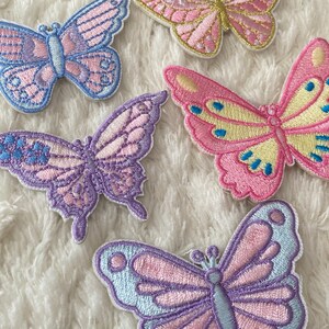 May include: Five embroidered butterfly patches in various pastel colours, including pink, purple, and blue. The butterflies are arranged on a white fluffy surface.