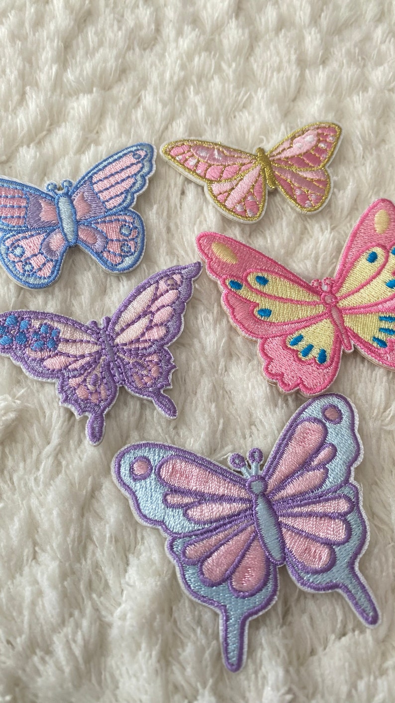 May include: Five embroidered butterfly patches in various pastel colours, including pink, purple, and blue. The butterflies are arranged on a white fluffy surface.