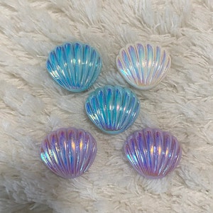 Puede incluir: Cinco cajas de alhajas con forma de concha de mar iridiscentes en varios tonos de azul y blanco. Las cajas están hechas de un material liso y brillante y tienen un diseño ligeramente en relieve.