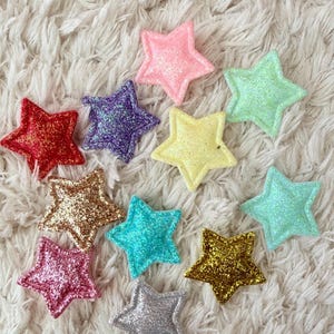 Pode incluir: Uma coleção de enfeites em forma de estrela brilhante em várias cores, incluindo vermelho, rosa, dourado, azul e verde. As estrelas estão espalhadas sobre uma superfície branca e fofa, criando um arranjo festivo e decorativo.