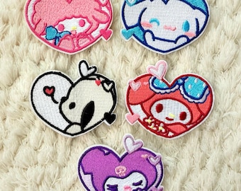 May include: Five heart-shaped embroidered patches featuring cartoon characters in various colors. The patches include pink, blue, black, red, and purple designs. Each patch has a white border and is set against a cream-colored, textured background.