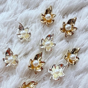May include: A collection of eight leaf-shaped buttons, each featuring a central white faux pearl. The buttons are finished in metallic gold or silver tones, with some displaying a smooth, reflective surface and others showing a pale yellow enamel.