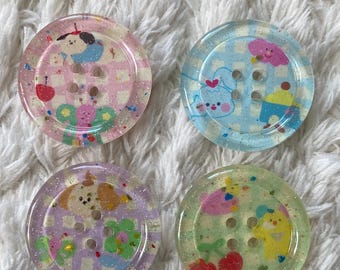 May include: Four colorful, round buttons with cartoon character designs. Each button has four holes for sewing and is decorated with glitter. The buttons feature pink, blue, purple, and green backgrounds with various cute illustrations.