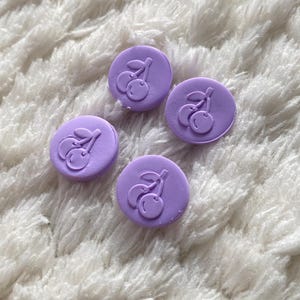 May include: Four small, round, lavender-colored buttons with a cherry design. The buttons are arranged on a white, fluffy surface. Each button features a raised image of two cherries and a stem.