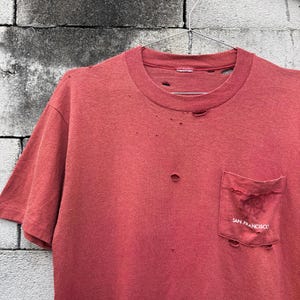 以下が含まれることがあります： クルーネックと胸ポケットが付いた、色あせた赤色の半袖Tシャツ。生地全体に複数の小さな穴やダメージが見られます。ポケットには薄い円形のグラフィックと「SAN FRANCISCO」という文字がプリントされています。