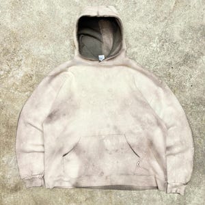 Könnte beinhalten: Beige Kapuzenpullover mit verblasstem Batik-Effekt. Der Hoodie hat eine Kängurutasche, eine Kapuze mit Kordelzug und gerippte Bündchen. Die Innenseite der Kapuze ist dunkelgrau.