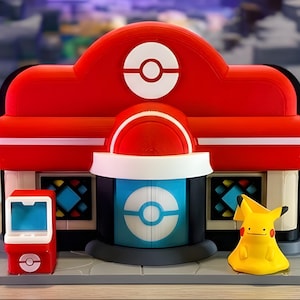 Puede incluir: Un modelo de edificio Centro Pokémon de plástico rojo y blanco con una puerta central azul con un emblema de Poké Ball blanco, un techo rojo con un icono de Poké Ball, una pequeña terminal roja y una figura amarilla de Pikachu.