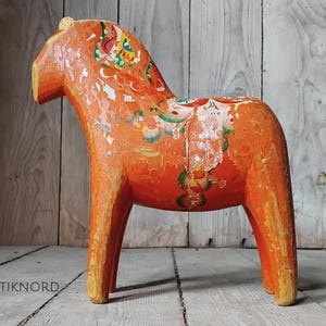 May include: A carved wooden horse figurine painted in a vibrant orange hue with distressed, weathered paint revealing wood grain. The horse features faded, multi-coloured floral patterns in green, yellow, and white across its body and mane.