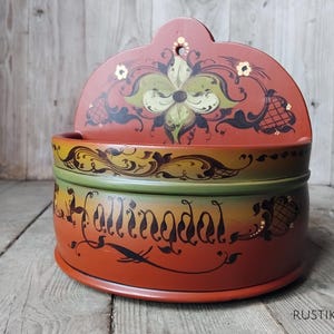 May include: A hand-painted, wooden wall-mounted container with a lid. The container is primarily red, with green and yellow accents. The lid features a floral design, and the side has the word "Hallingdal" in script.