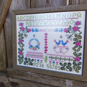 May include: Framed cross-stitch sampler with a floral and bird design. The embroidery features pink roses, blue birds, and the year "1973." The sampler includes a wreath, hearts, and a border of green leaves and flowers. The frame is a light brown color.