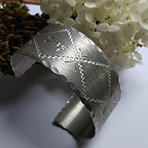 Può includere: Un bracciale a polsino argentato con superficie strutturata e design geometrico. Il bracciale presenta forme a diamante con dettagli incisi e un bordo smerlato. Fiori bianchi e verde sullo sfondo.