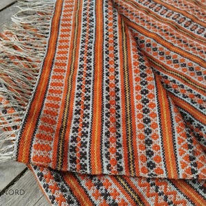 Könnte beinhalten: Gewebter Textil mit einem sich wiederholenden Muster aus orangefarbenen, schwarzen und weißen vertikalen Streifen und floralen Motiven. Die Kanten sind mit einem Fransen versehen. Der Textil ist gefaltet und zeigt das Muster und die Textur.