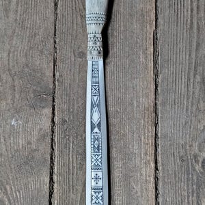May include: A decorative, hand-carved knife with a light gray handle and blade. The handle is shaped like an antler and features intricate geometric patterns. The blade is long and slender, also adorned with detailed designs. The knife rests on a wooden surface.