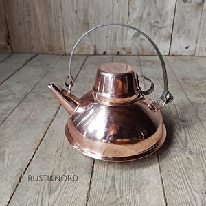 May include: A copper kettle with a spout and a handle. The kettle has a shiny, reflective surface and a lid. The handle is made of metal and is attached to the kettle with metal loops. The kettle is sitting on a wooden surface.
