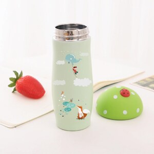 May include: A green stainless steel water bottle with a whimsical design featuring a fox, a boy, and a whale. The bottle has a green lid with a ladybug on top.