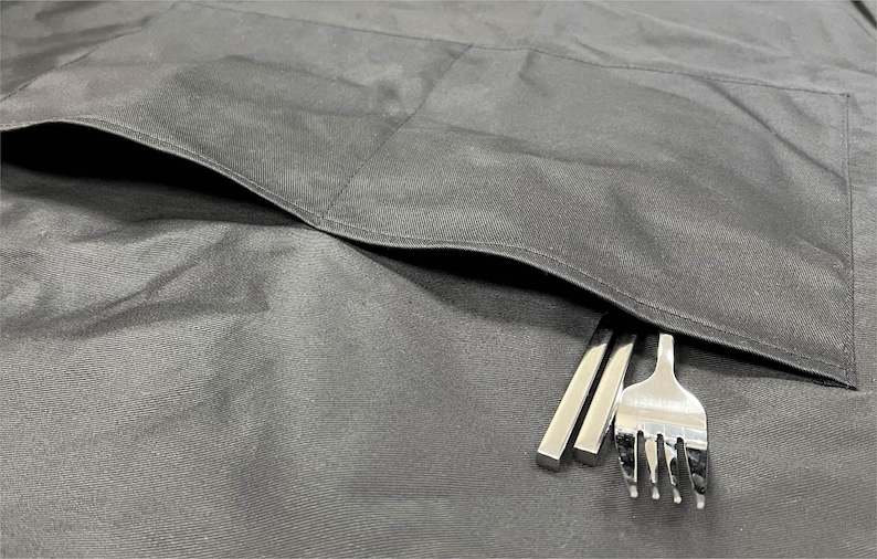 May include: A black fabric apron with a large pocket on the front. Two silver chopsticks and a silver fork are sticking out of the pocket.