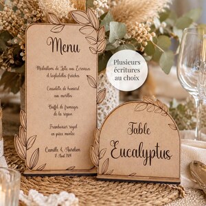 Peut inclure: Un menu rectangulaire en bois et un numéro de table semi-circulaire en bois, tous deux ornés de motifs de feuilles gravés au laser. Le menu affiche le texte "Menu", "Camille & Aurelien", "17 Aout 2024" et le panneau de table indique "Table Eucalyptus".