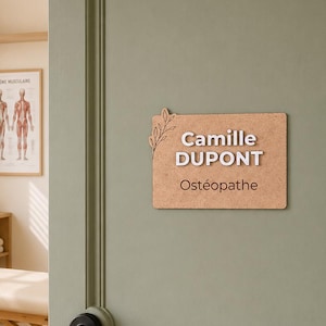 Op de afbeelding: Een rechthoekig lichtbruin houten bordje bevestigd op een saliegroene deur. Het bordje bevat de witte verhoogde tekst Camille DUPONT en de bruine tekst Ostéopathe, met een decoratief uitgesneden bladdetail aan de linkerzijde van het bord.