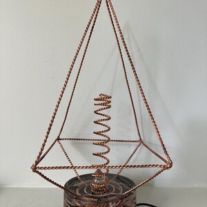 Puede incluir: Una escultura en forma de pirámide hecha de alambre de cobre trenzado, con una bobina central vertical de alambre de cobre. La estructura está montada sobre una base circular de resina oscura que contiene alambre de cobre y piedras oscuras.