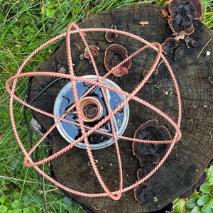 May include: A copper-coloured wire sculpture formed into a five-pointed star shape, positioned over a clear glass circular container filled with a dark liquid. The sculpture features a twisted metal texture and sits centred above the glass vessel.