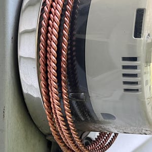 May include: Three strands of twisted copper wire wrapped around the circular side of a grey utility meter. The copper wire has a metallic, reddish-brown finish and a repeating spiral texture along its entire length, contrasting with the smooth grey casing.