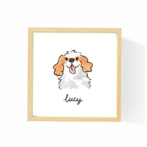 May include: A framed watercolor illustration of a Cavalier King Charles Spaniel dog with orange and white fur. The dog is smiling with its tongue out. The name "lucy" is written in cursive below the dog.
