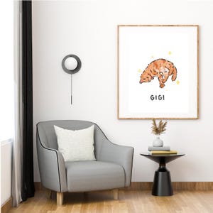 May include: A framed art print featuring a cartoon illustration of a ginger cat with the name "GIGI" below. The artwork is displayed on a white wall above a gray armchair with a white pillow. A small black side table holds books and a vase.