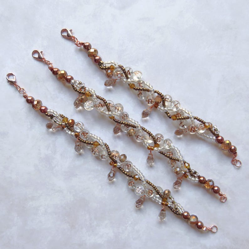 May include: Three beaded bracelets with copper-colored clasps and accents. The bracelets feature clear and amber-colored beads, with some spelling out letters. The design includes dangling teardrop-shaped beads.