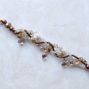 May include: A beaded bracelet with clear, brown, and gold beads. The bracelet has a copper-colored clasp and accents. Some beads spell out the word "CREATE". The bracelet is on a white surface.
