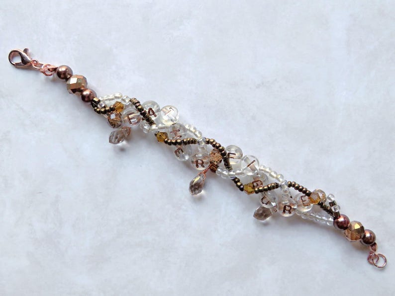 May include: A beaded bracelet with clear, brown, and gold beads. The bracelet has a copper-colored clasp and accents. Some beads spell out the word "CREATE". The bracelet is on a white surface.