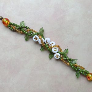 May include: A beaded bracelet with the name "WILLOW" spelled out in white letter beads. The bracelet features green and gold beads, with small green leaf accents. Orange and yellow beads are at each end.
