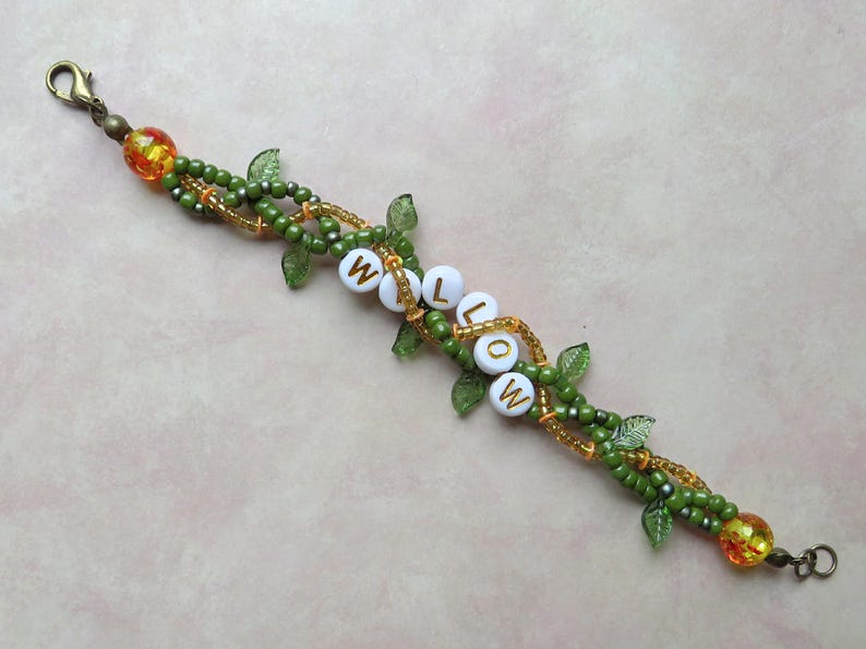 May include: A beaded bracelet with the name "WILLOW" spelled out in white letter beads. The bracelet features green and gold beads, with small green leaf accents. Orange and yellow beads are at each end.