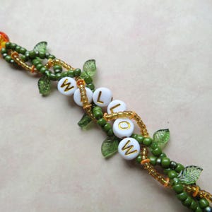 May include: A beaded bracelet with the word "WILLOW" spelled out in white letter beads. The bracelet features green beads, gold accents, and leaf-shaped charms. The clasp is gold and orange.