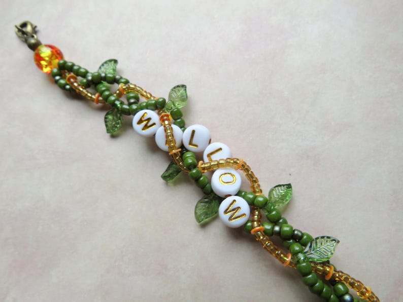 May include: A beaded bracelet with the word "WILLOW" spelled out in white letter beads. The bracelet features green beads, gold accents, and leaf-shaped charms. The clasp is gold and orange.