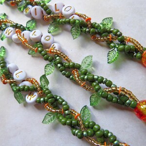 May include: Close-up of beaded bracelets with the word "WILLOW" spelled out in white letter beads. The bracelets feature green beads, gold accents, and clear leaf-shaped beads. Each bracelet ends with a decorative yellow and red bead.
