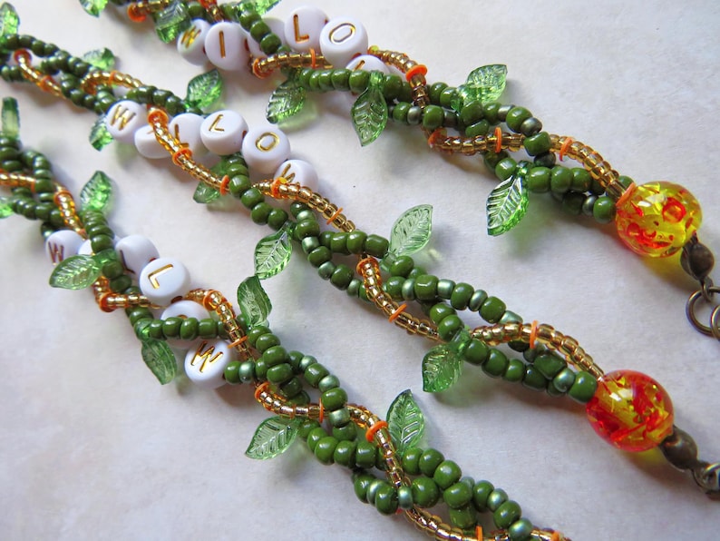 May include: Close-up of beaded bracelets with the word "WILLOW" spelled out in white letter beads. The bracelets feature green beads, gold accents, and clear leaf-shaped beads. Each bracelet ends with a decorative yellow and red bead.