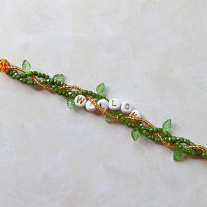 May include: A beaded bracelet with the word "WILLOW" spelled out in white letter beads. The bracelet features green and gold beads, with small green leaf accents. The ends have decorative orange and gold beads.