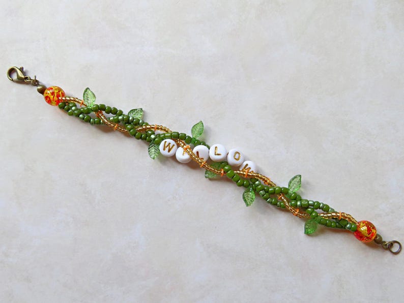 May include: A beaded bracelet with the word "WILLOW" spelled out in white letter beads. The bracelet features green and gold beads, with small green leaf accents. The ends have decorative orange and gold beads.