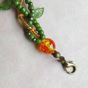 May include: A beaded keychain with a nature theme. The keychain features green beads, gold accents, and a large, round, orange and yellow glass bead. The clasp is a bronze color, and there are green leaf accents.